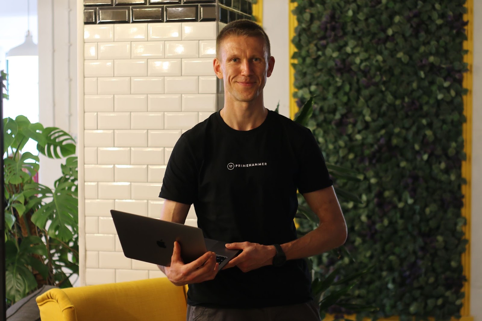 Zbynek poses for a photo standing up on an indoor background, with a laptop on his arm and a black primehammer T-shirt.