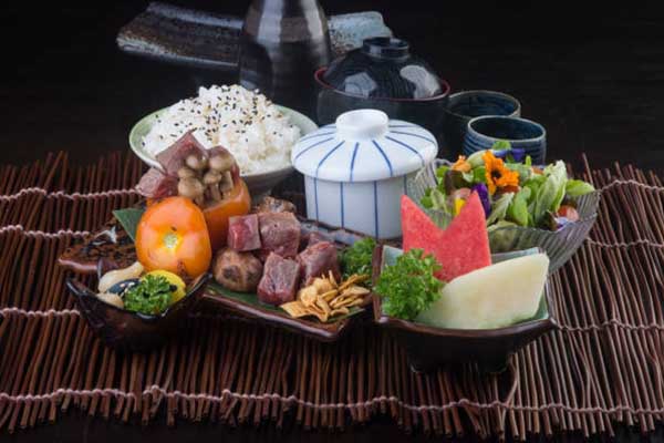  A meal arrangement featuring meat, rice sprinkled with sesame, stuffed tomatoes, a salad with edible flowers, and assorted condiments, presented on a bamboo mat.