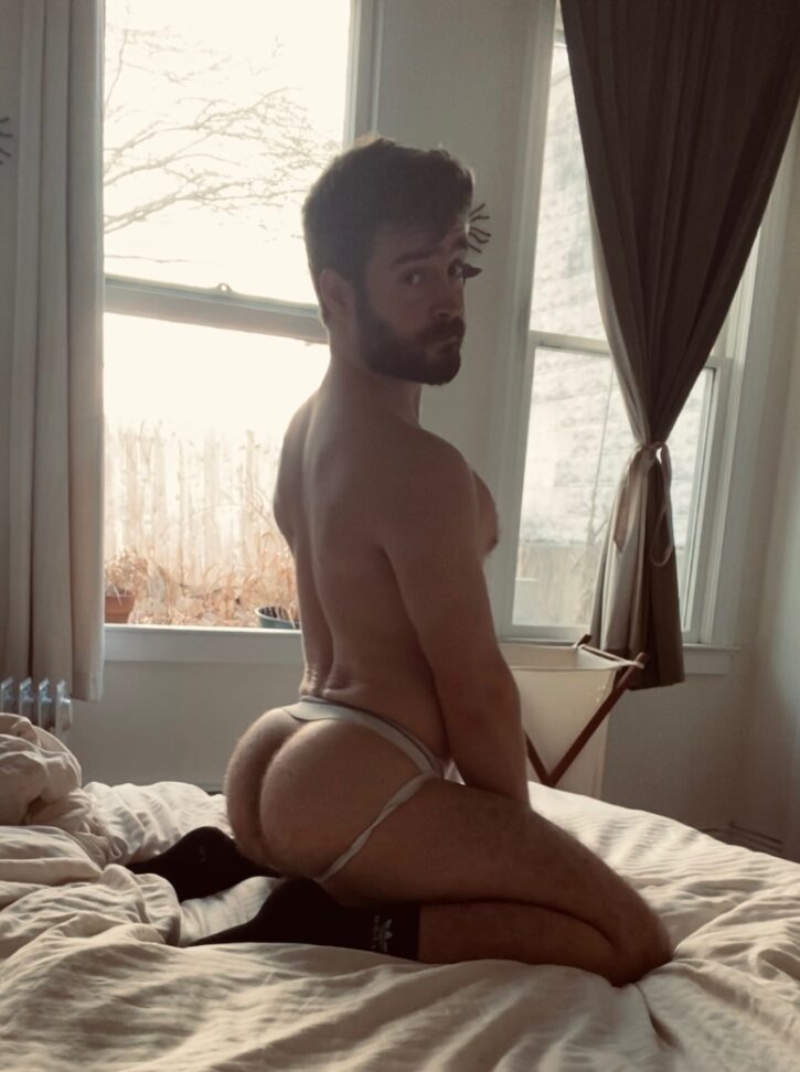 Forest Cliff kneeling naked on the bed in a white jockstrap showing off his hairy gay bubble butt while looking over his shoulder