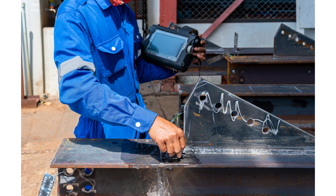 With a screen in his left hand, and a sensor in his right, an inspector dressed in a blue jumpsuit uniform uses a tool to measure  a metal surface, looking for any defects in the metal, such as a bowed profile.