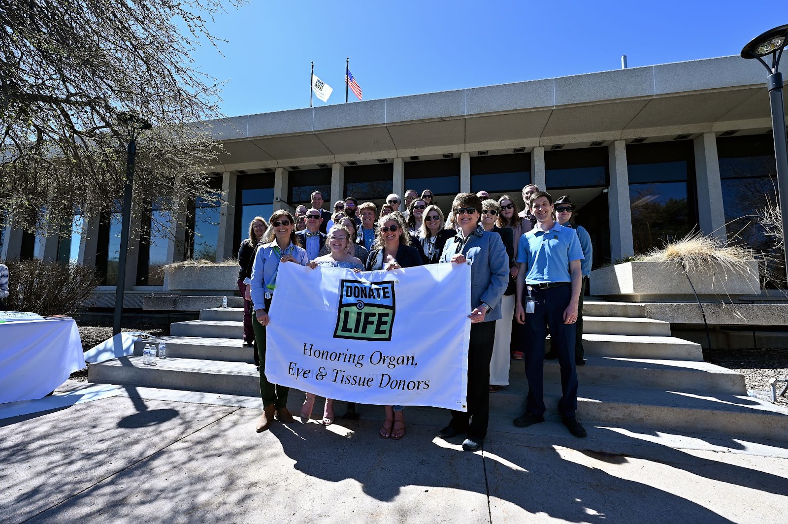 Sixteen-year-old Lella Young, Colorado Division of Motor Vehicles Senior Director Electra Bustle and Donate Life Colorado CEO Jennifer Prinz pose with about 20 people for a photo Thursday, April 4, 2024, after a flag-raising ceremony at DMV headquarters in Lakewood.