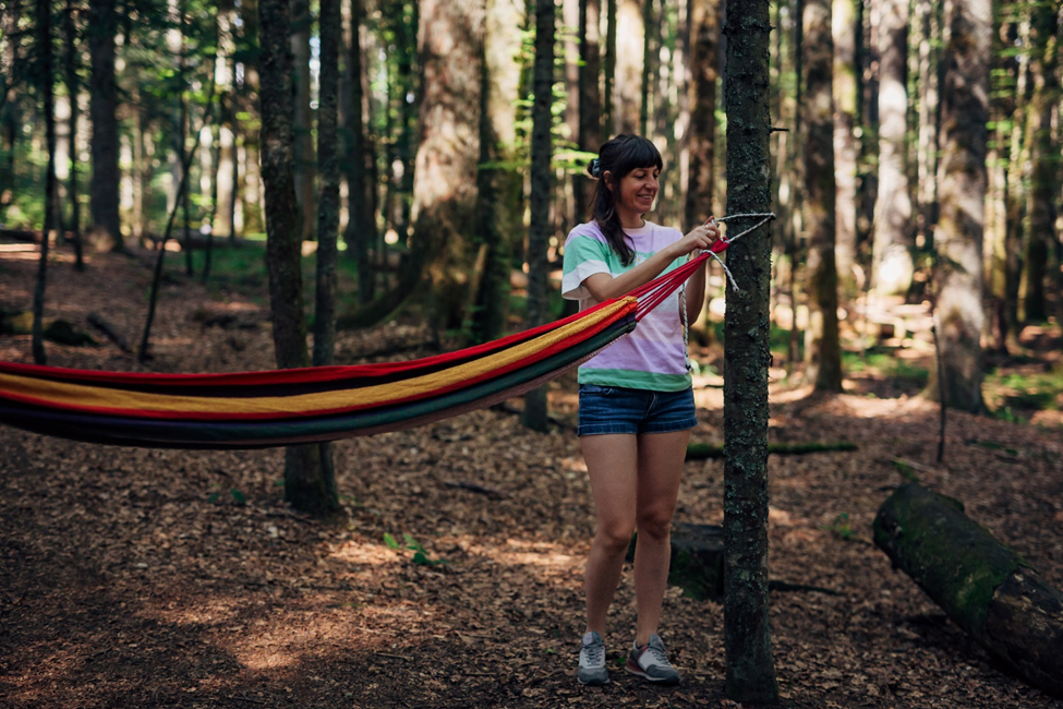 A young woman hangs one end of a hammock to the end of a tree. 