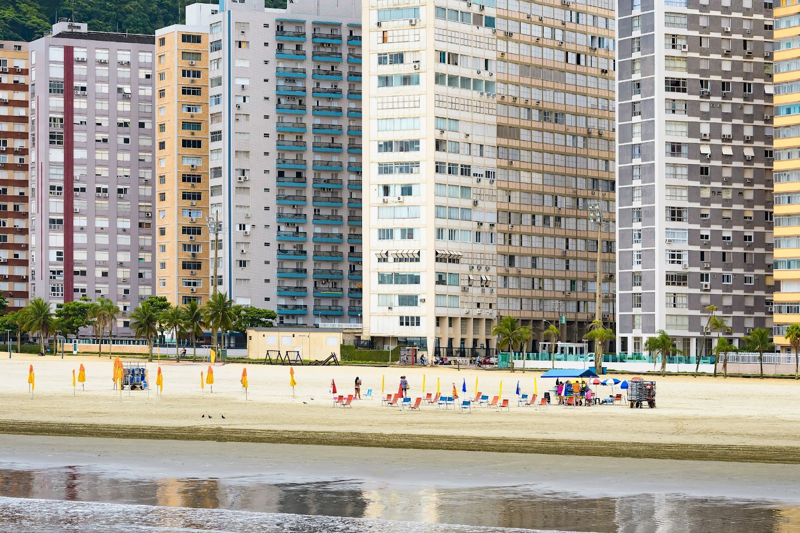 Faixa de areia de uma das praias de Santos com alguns banhistas em cadeiras de praia de frente para o mar. Em segundo plano, atrás da faixa de areia, diversos prédios de cores diferentes.