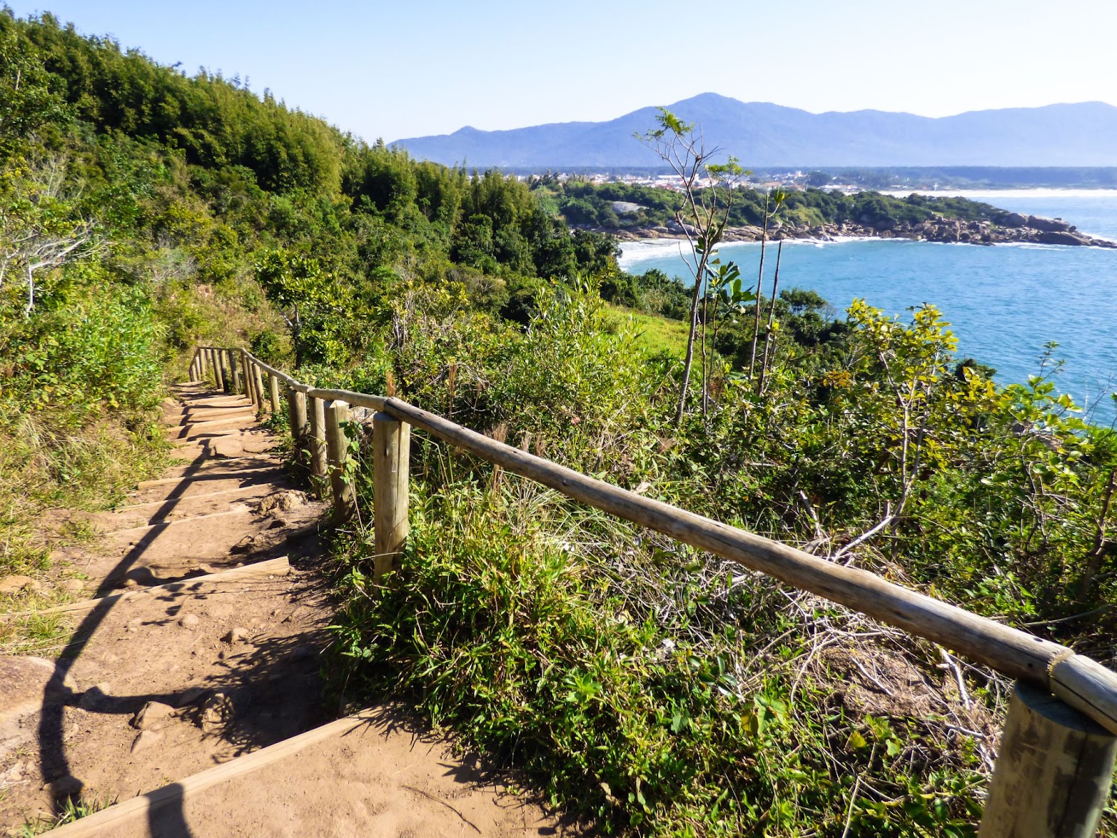 Escadaria que leva até as piscinas naturais da Barra da Lagoa, em Florianópolis. Os degraus de terra batida estão cercados por vegetação verde. Ao fundo, aparecem o mar azul e as montanhas.
