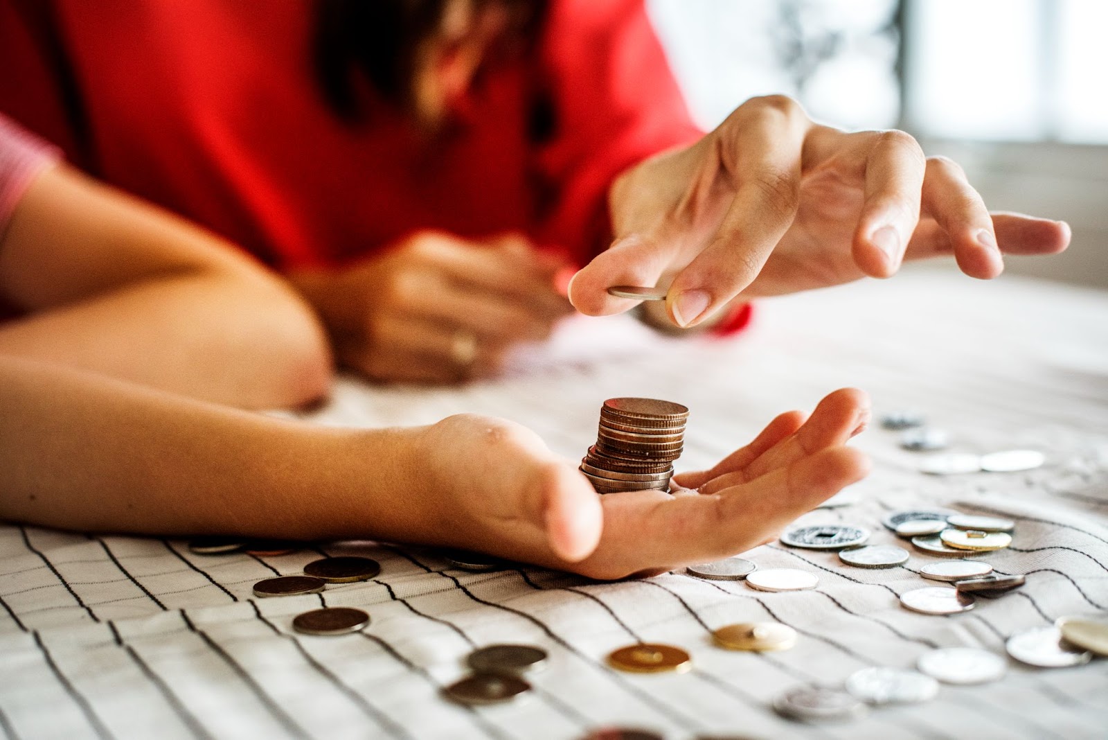 Imagem de uma pilha de moedas sendo colocadas na palma da mão de alguém. Ao redor das mãos, há diversas moedas espalhadas.