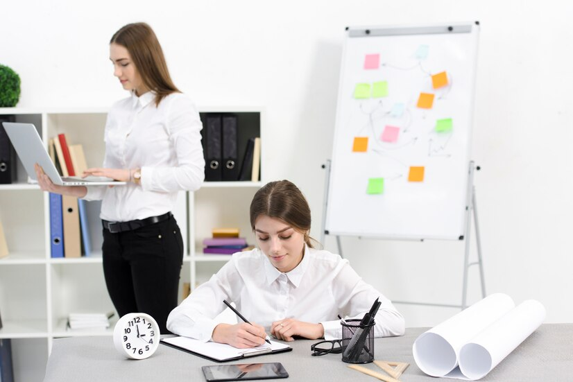 businesswoman-writing-notes-clipboard-with-her-female-colleague-using-digital-tablet-office