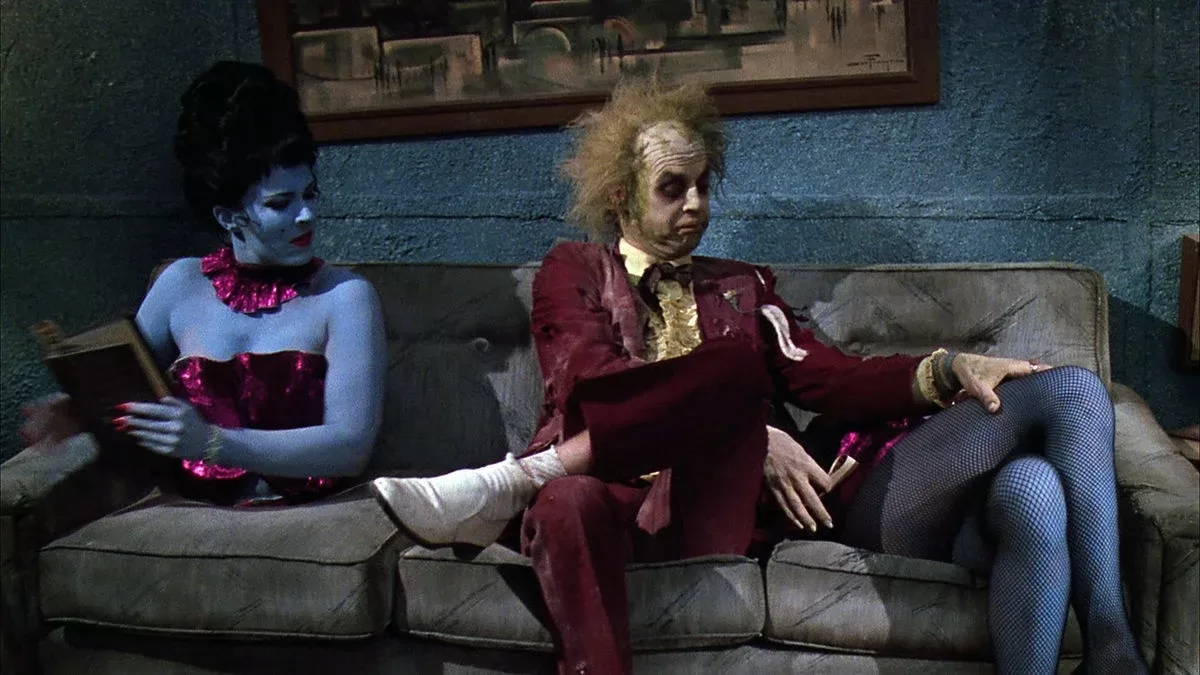 Beetlejuice (Michael Keaton) comparte en un sofá con una señorita bastante peculiar: su torso está a un lado (izquierda) y sus piernas cruzadas al otro (derecha).
