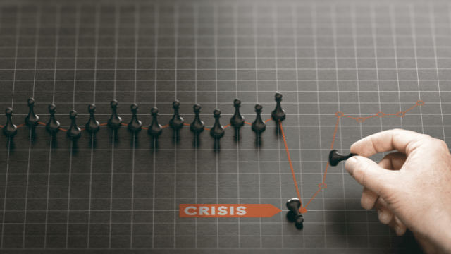 A hand holding a chess piece in front of a downward trending graph. Text at the top reads “CRISIS”.