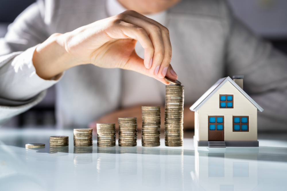 Foto de uma pessoa com camisa cinza, que aparece as mãos empilhando várias moedas e uma miniatura de casa ao lado representando um consórcio imobiliário.