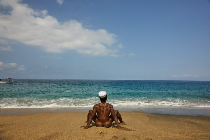 Jordan Jameson sitting naked on the beach towards the ocean wearing a sailors hat