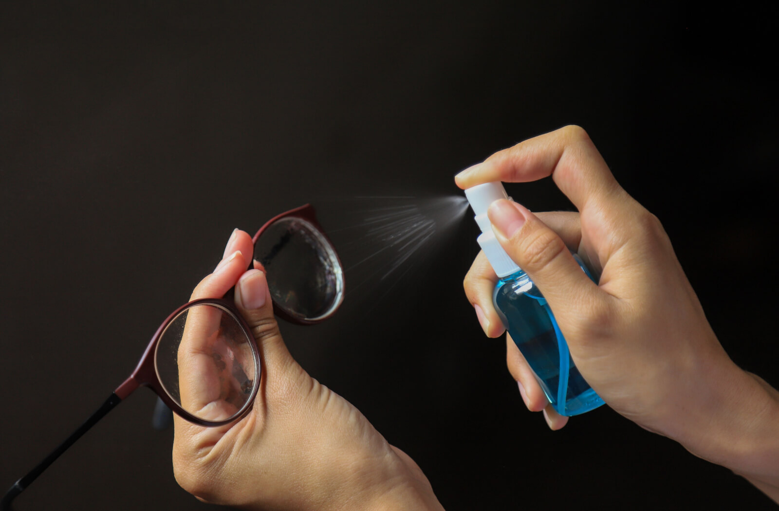 A close-up of hands spraying a cleaning solution unto a pair of eyeglasses.