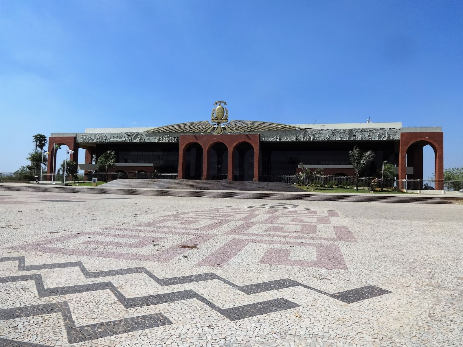 Foto tirada da fachada do Palácio Araguaia. No chão, pedras portuguesas que formam desenhos. Céu limpo e azul