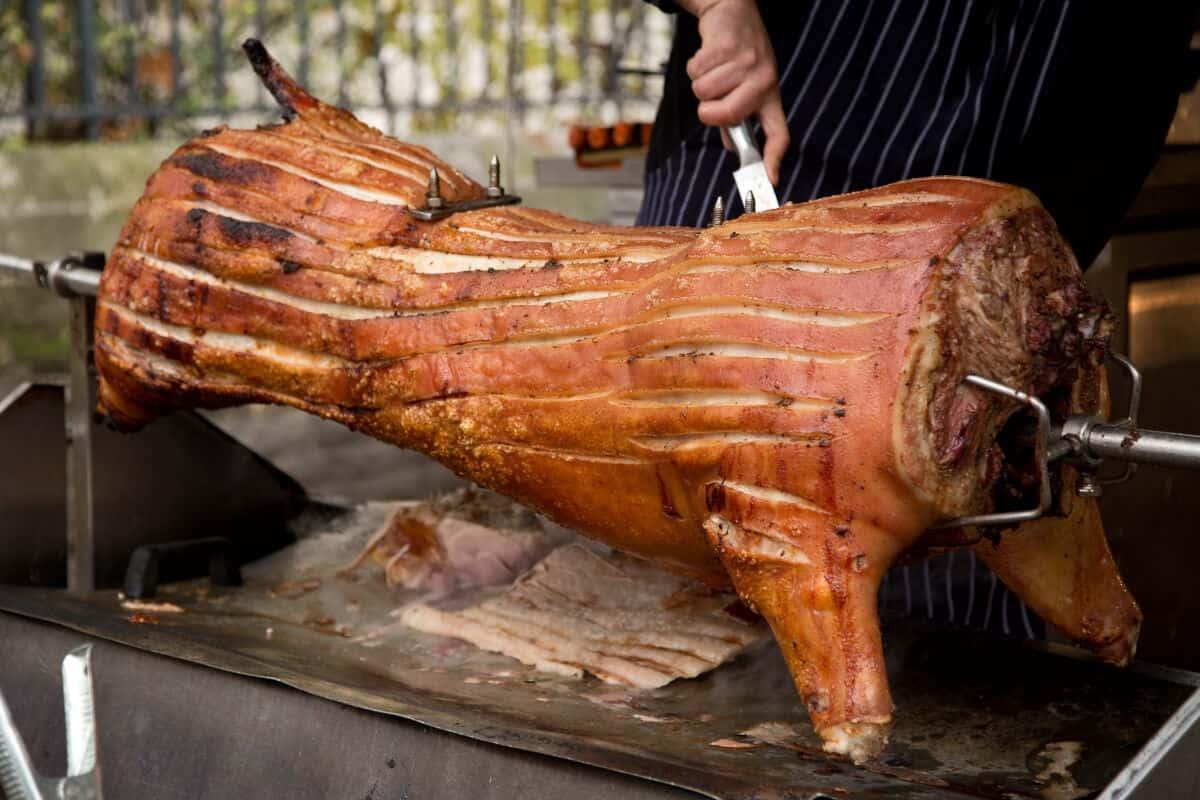 Un porc întreg pe rotisor, cu pielea tăiată și un bărbat pe cale să-l felieze