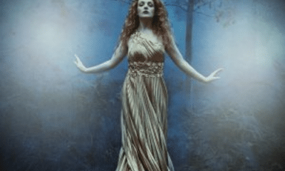 Badb is wearing a gold dress with her hands stretched out She is in a misty woods. 