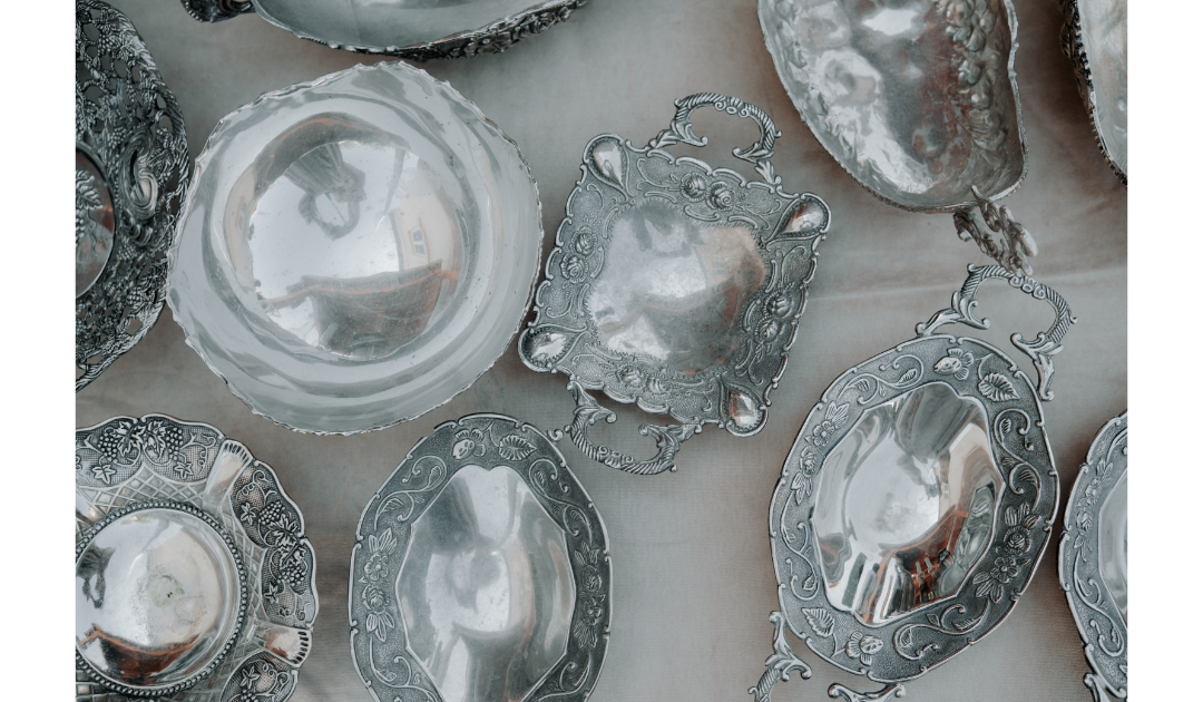 Silver platters of different sizes, shapes, and levels of shine are laid out amongst a cloth. In the reflections, there are distortions of someone taking a picture. 