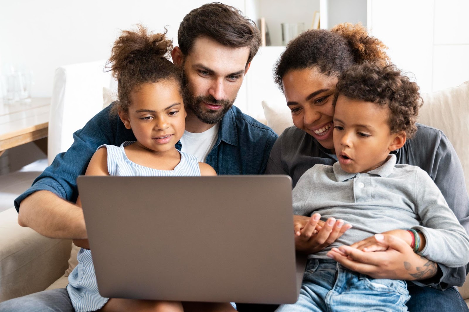 Imagem de uma família olhando a tela de um notebook. Há um homem e uma mulher sentados em um sofá e, em seus colos, há duas crianças pequenas. Os adultos sorriem.