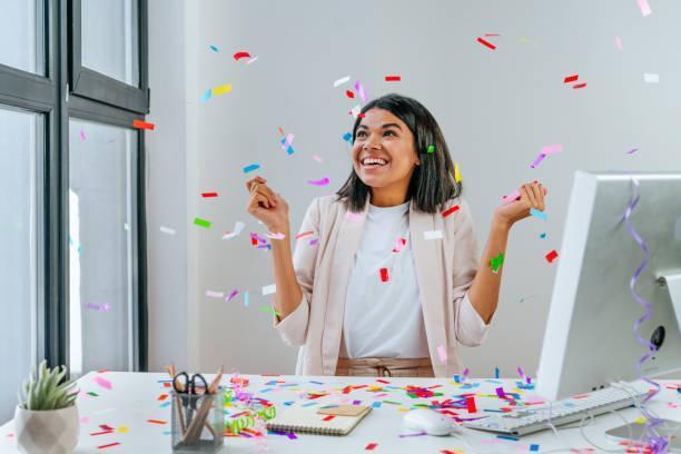 Young business woman having fun time catching confetti Young business woman having fun time catching confetti sitting at the desk in the office. Party time on the work place. Selective focus. business holiday greetings cards stock pictures, royalty-free photos & images