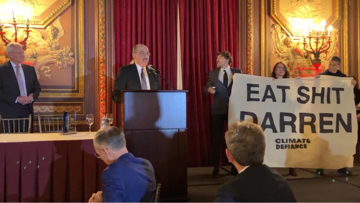 The CEO addresses the room but beside him stand three activists holding a banner that says Eat Shit Darren.