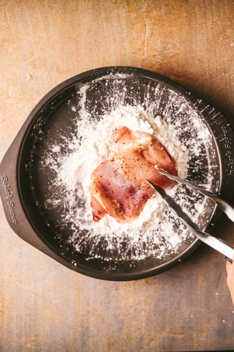 A person is sprinkling flour on top of chicken in a pan.