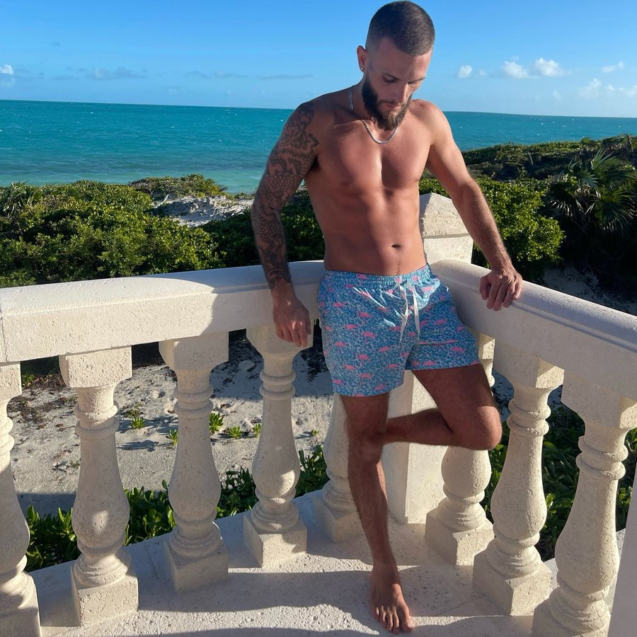 Tony Salas posing in a blue swim shorts on the balcony in a tropical location taking a photo for onlyfans content