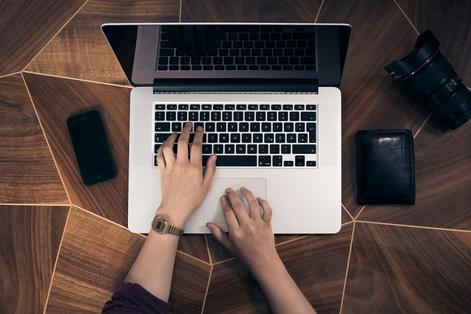 A person working on a desk in a laptop, phone, camera and wallet on the side