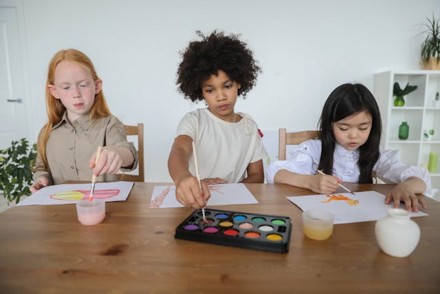 Three children engaged in painting activities at a wooden table with art supplies