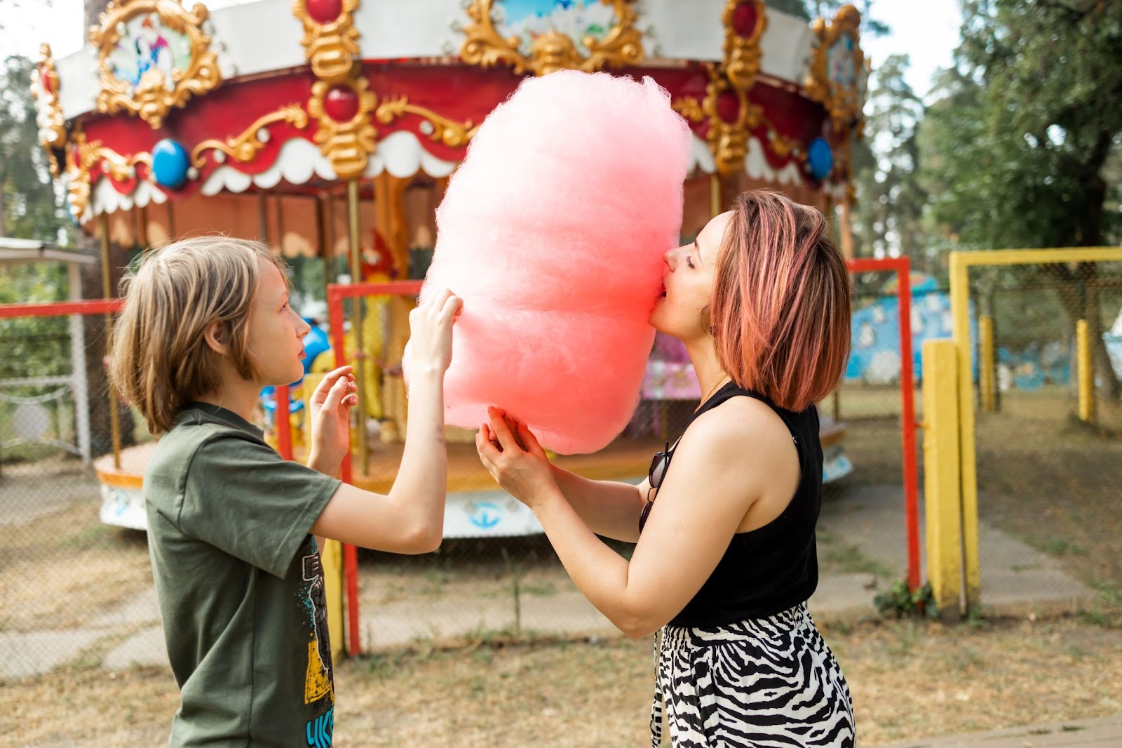 Mom and son enjoy pink cotton candy in front of the carnival carousel.