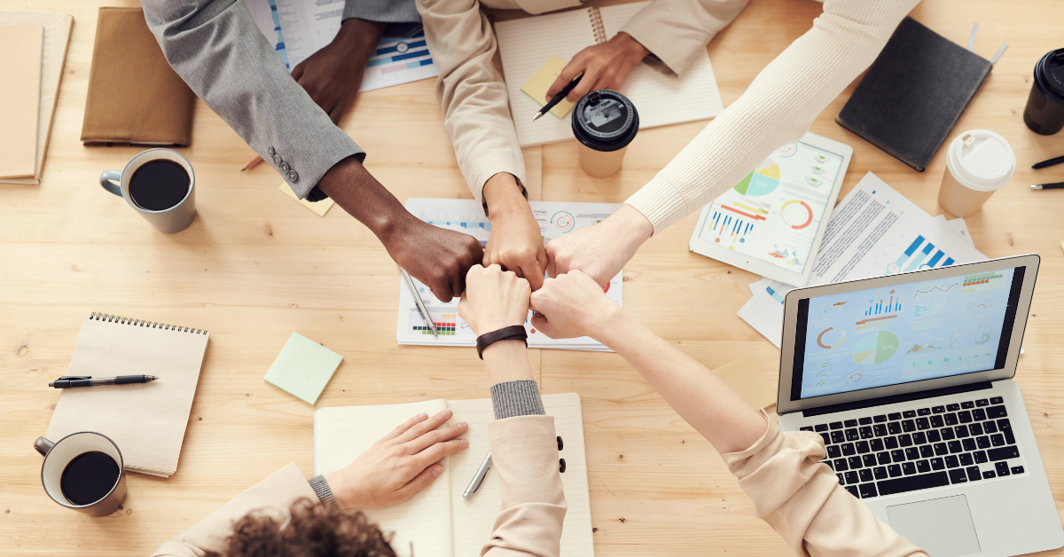 Top view of team members fist bumping over a table with charts, notebooks, and coffee cups.