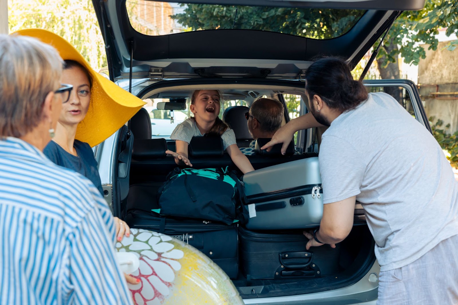 Imagem de uma família se preparando para viajar. A fotografia é tirada de frente para um porta-malas aberto, com diversas malas sendo posicionadas dentro do veículo. É possível ver 5 pessoas, sendo uma delas uma criança no banco de trás.