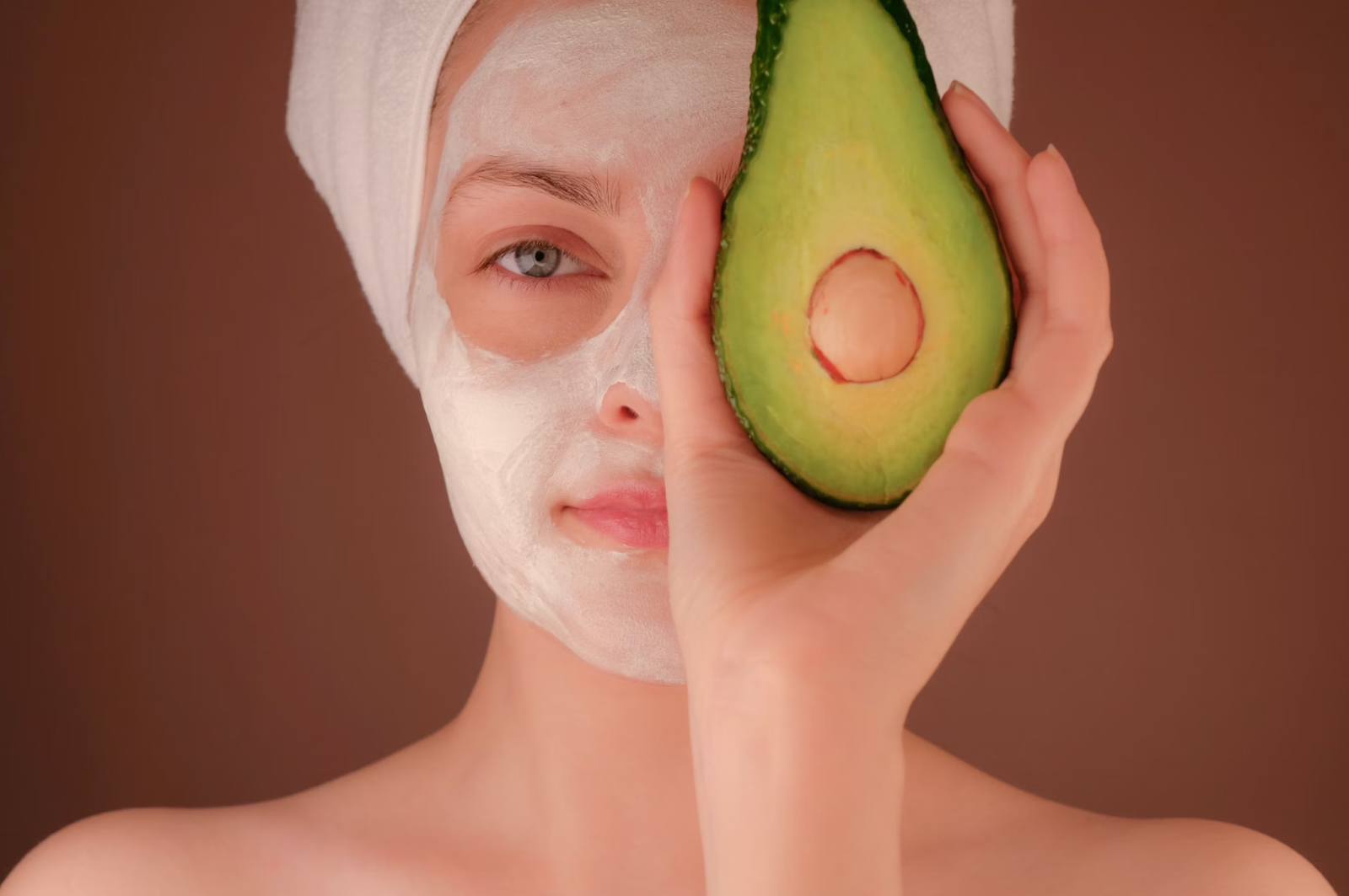 woman with white face mask applied holding up half an avocado