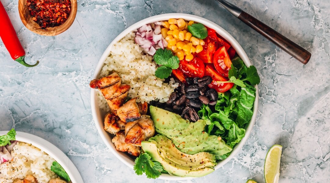 bowl with chicken, quinoa, onion, chickpeas, spinach, tomatoes, beans, and avocados on a gray countertop