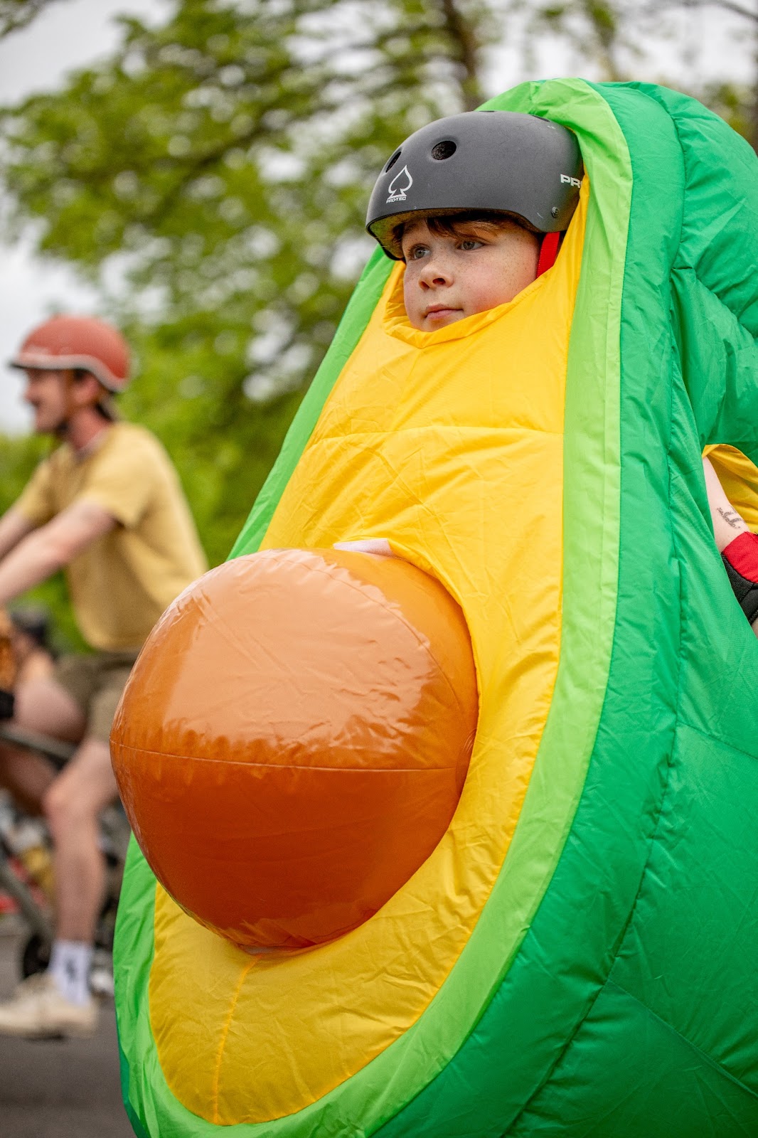 A little kid in a bike helmet and an inflatable avocado costume with a big pit on his tummy at the Ladd's 500 relay in Portland, in April of 2024.