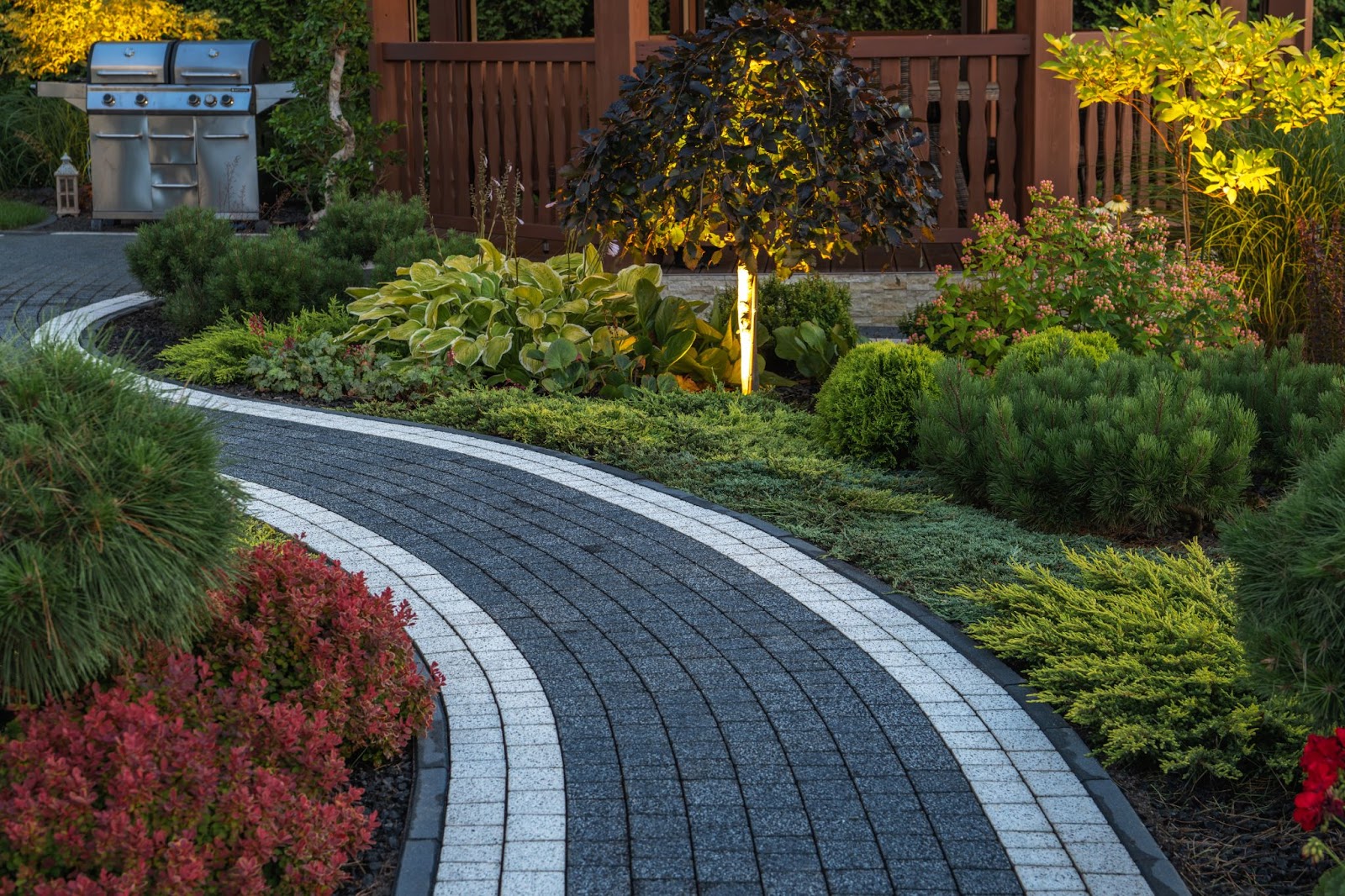 A well-paved concrete brick garden pathway leading to a brown-fenced porch, surrounded by a lush garden filled with variously colored plants.