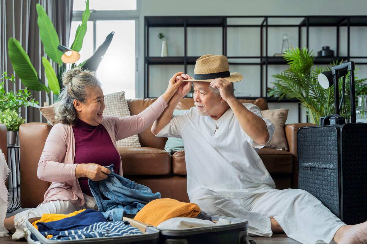 An older couple sits on the floor, laughing with each other, and packs a suitcase for vacation.