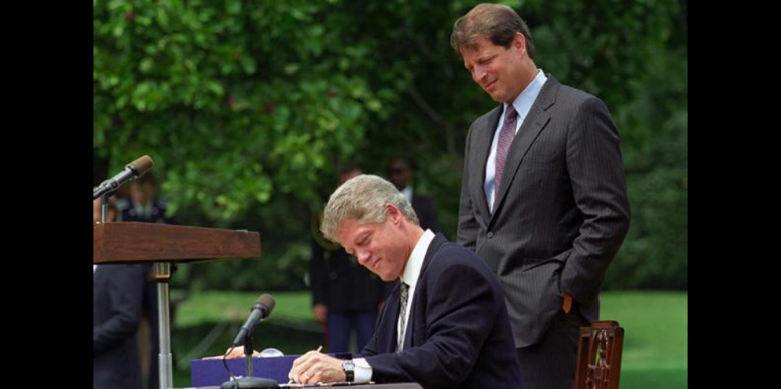 Bill Clinton signs a bill into law while Al Gore looks on. They are outdoors. 
