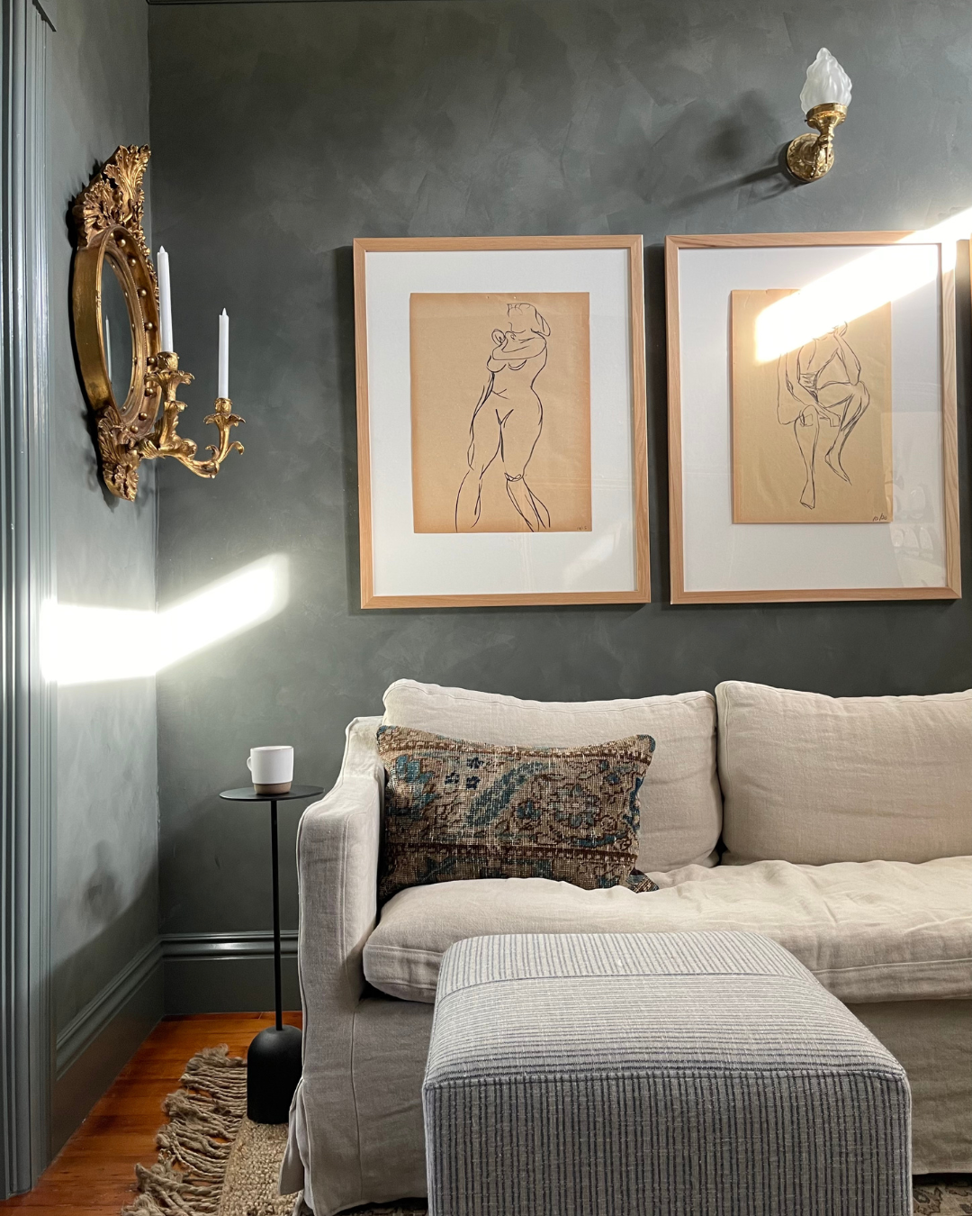 Kim Lucian's living room with gray painted walls, a pair of man and women pencil sketched paintings, a gilded Girandole mirror, a comfortable linen sofa and ottoman, and a modern black steel side table. The space harmonizes moody colors and ornate details.