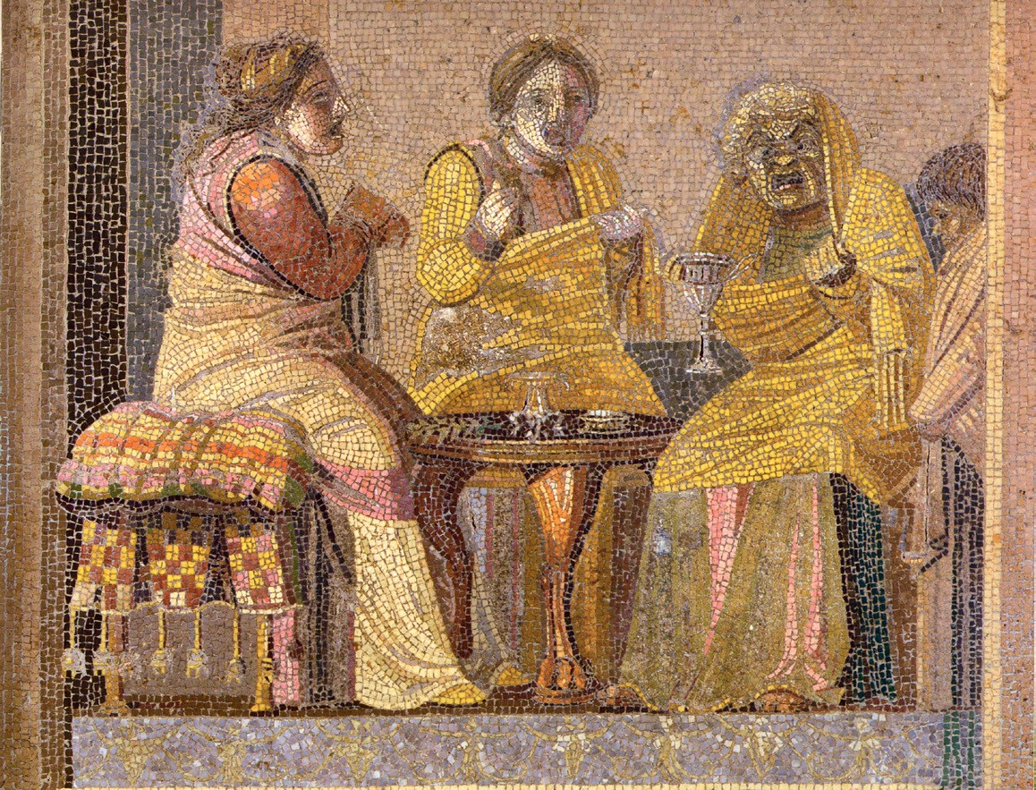 A mosaic of three women sitting at a small table. One is older and two are younger. They all appear to be wearing full face masks along with ancient Roman clothing.  