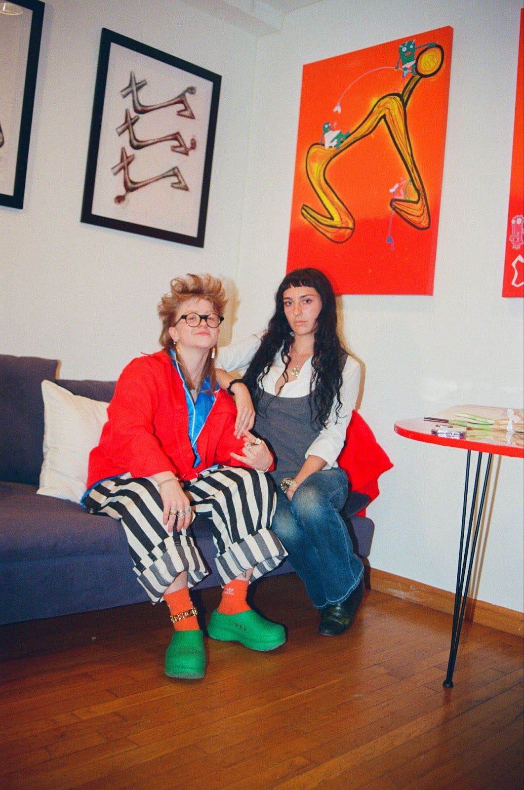 From left to right, a blonde girl with round glasses, a red jacket, and black and white striped pants sits next to a girl with long black hair and a white button up shirt on.