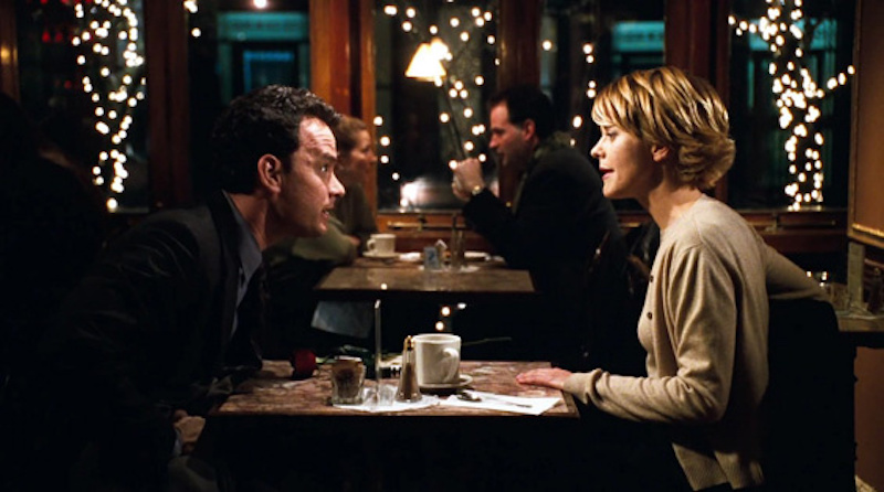 Joe Fox (Tom Hanks) and Kathleen Kelly (Meg Ryan) sit face to face in a cozy diner, sharing a moment of connection, akin to the heartwarming ambiance found in the best comfort Christmas films. Their eyes meet with a mix of curiosity and anticipation as they engage in conversation, unaware of each other's true identity as online pen pals.