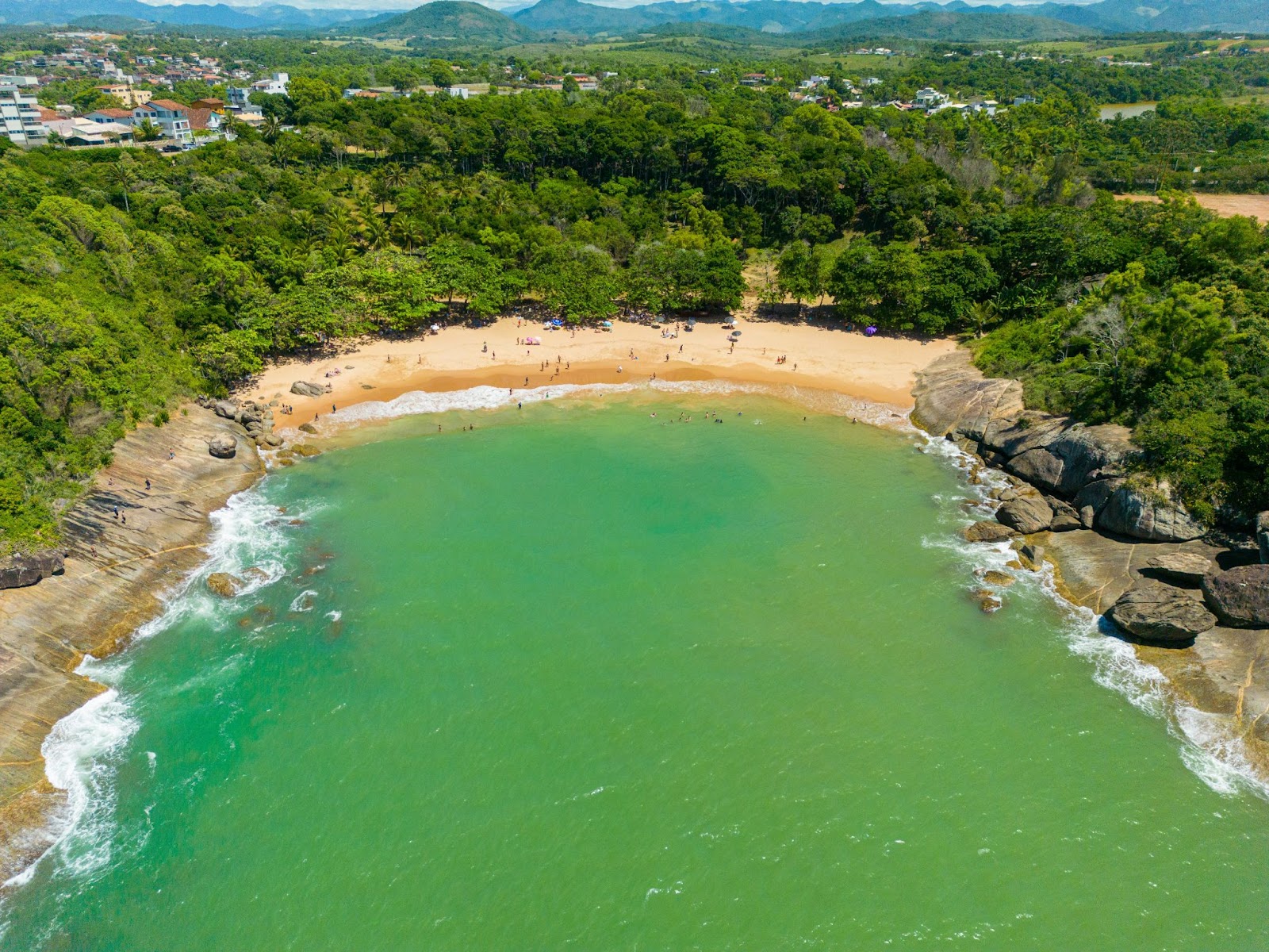 Vista aérea panorâmica das praias de Bacutia, Peracanga e dos Padres. Faixa de areia em formato de concha levemente centralizado na imagem, com mar esverdeado à sua frente.