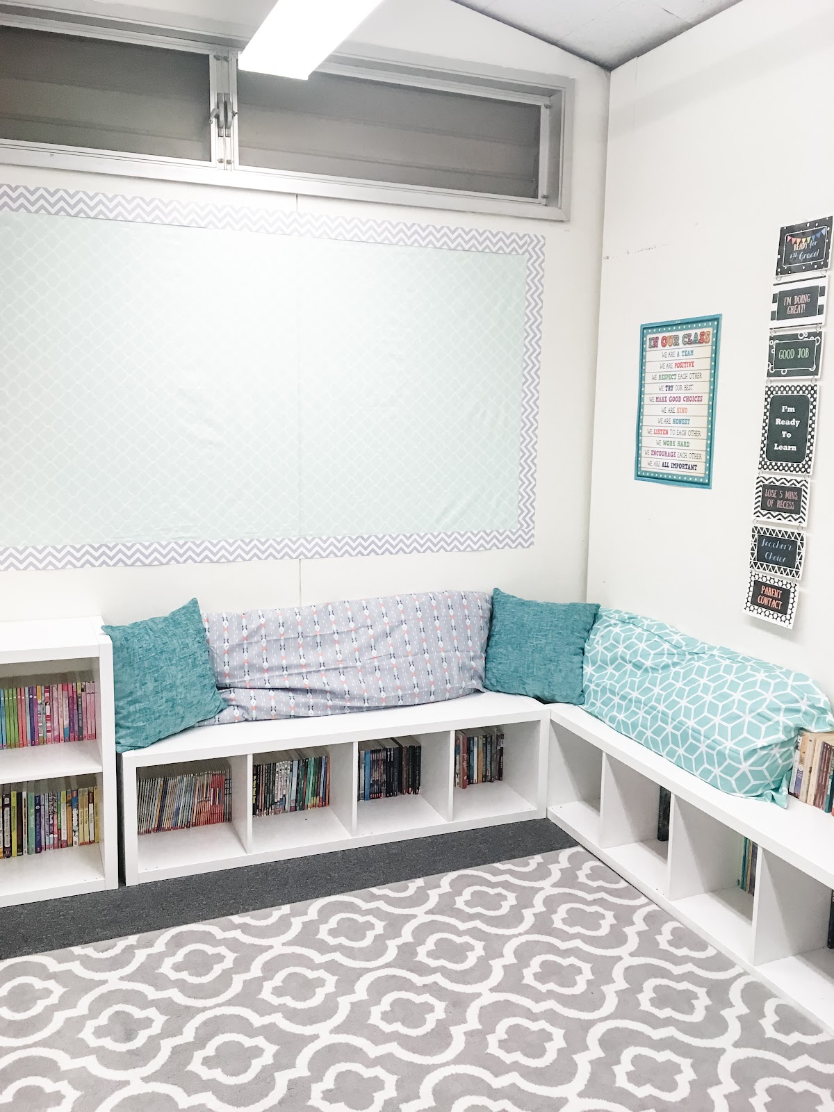 This image shows a closeup of a reading nook with low bookshelves that double as a bench. There are teal and gray pillows on top of the bench. 