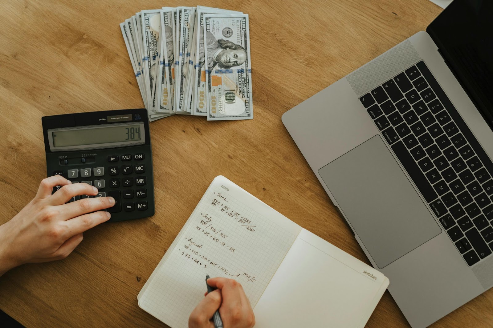 A person using a calculator and writing in a notebook, with stacks of hundred-dollar bills and a laptop on the desk.