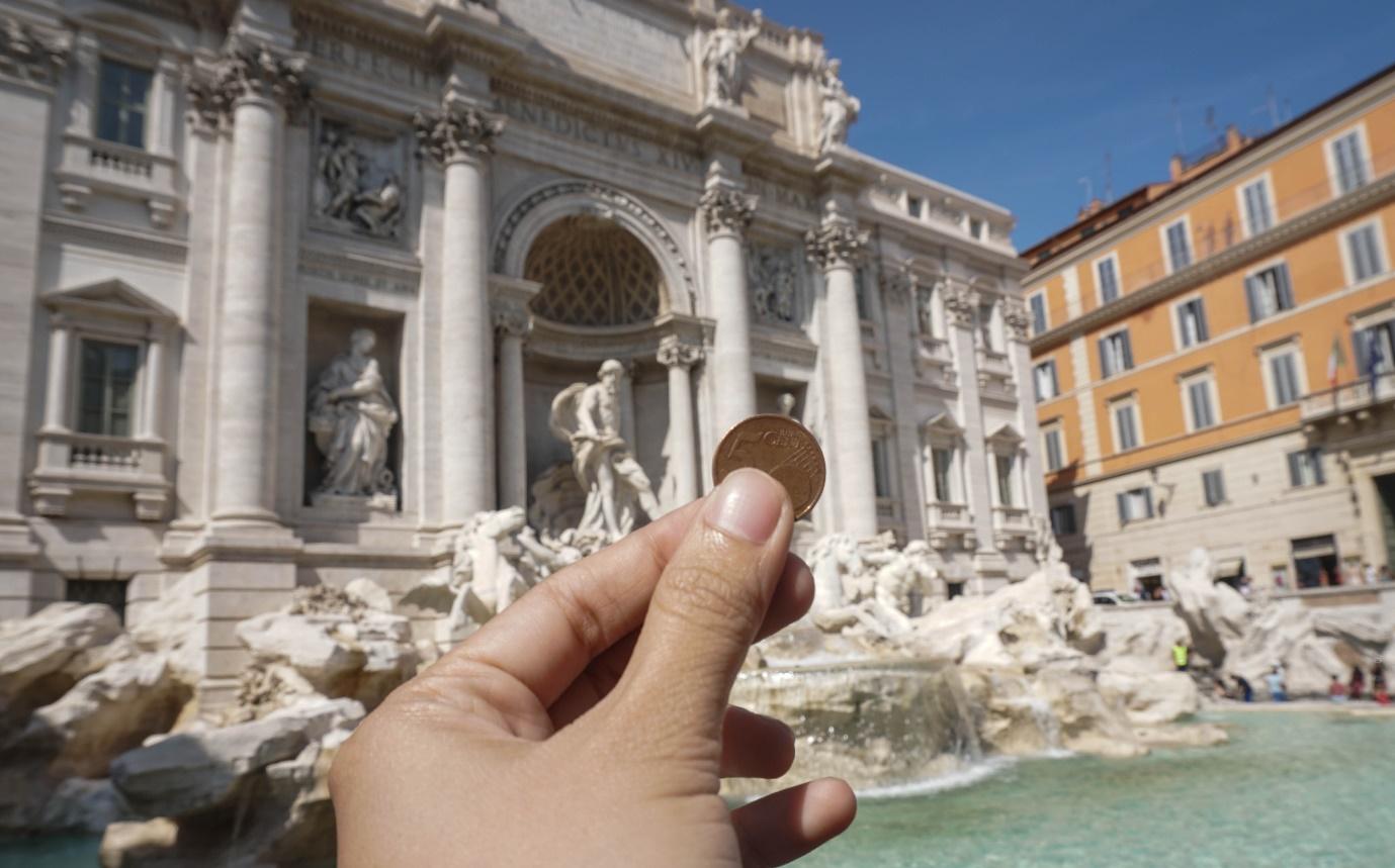 A hand holding a coin in front of a fountain with Trevi Fountain in the background

Description automatically generated