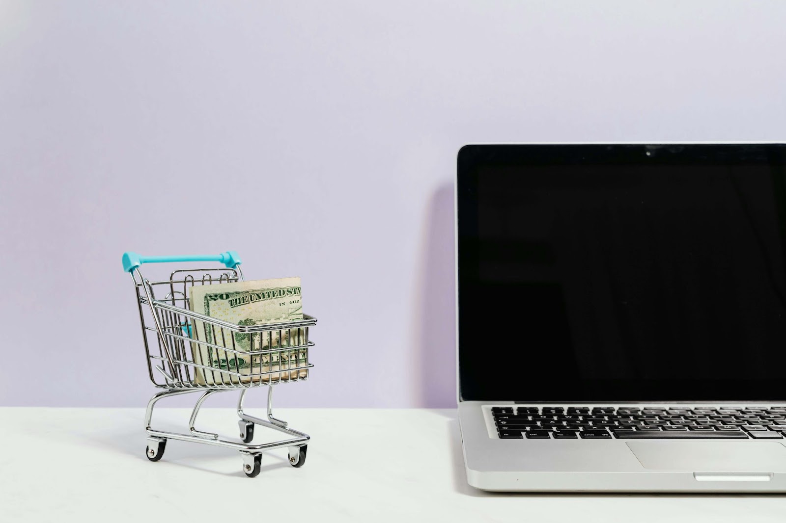 Carrito de compras en miniatura con billetes de dólar junto a una laptop, representando la facilidad de ventas en WhatsApp