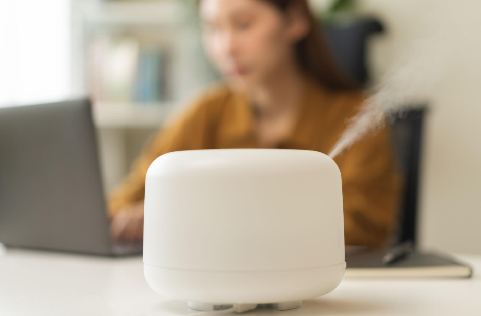 Close-up of a humidifier on a desk. In the blurred background, a woman sits and works at a laptop.