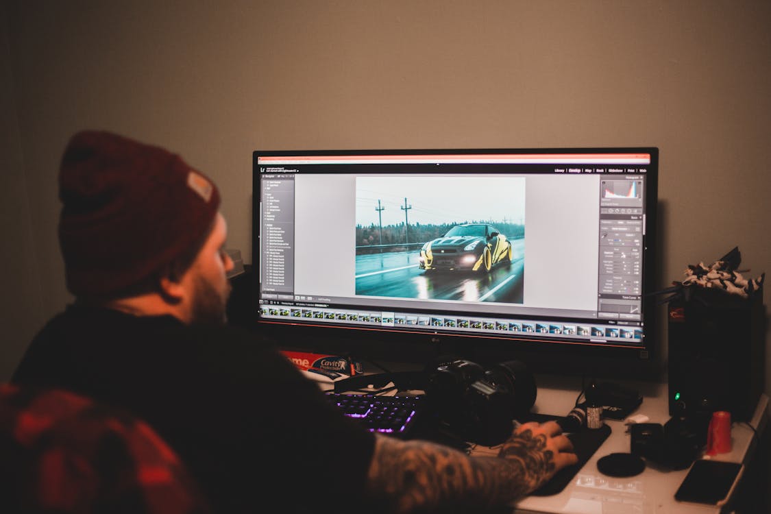 Free Unrecognizable tattooed male sitting at table and working on computer with montage video of sport car Stock Photo