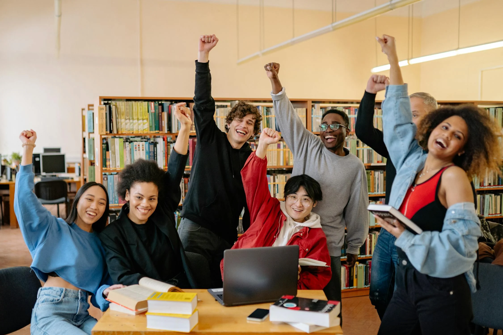 A group of seven students sitting in a library gathered around a table, holding their fists up and smiling - 10 Extracurricular Activities to Boost Your College Application