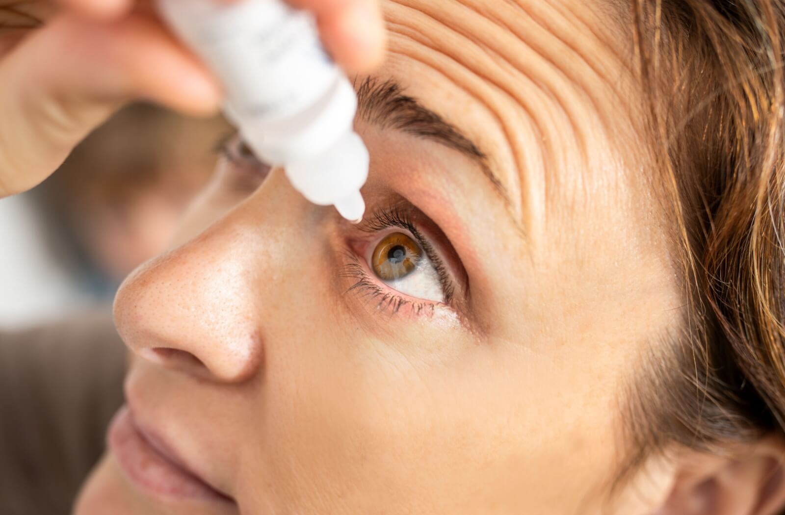 A close-up of a woman applying artificial tears to her left eye.