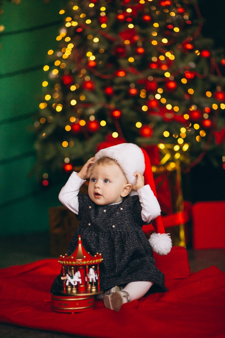 A baby sitting by the Christmas tree with her toy.