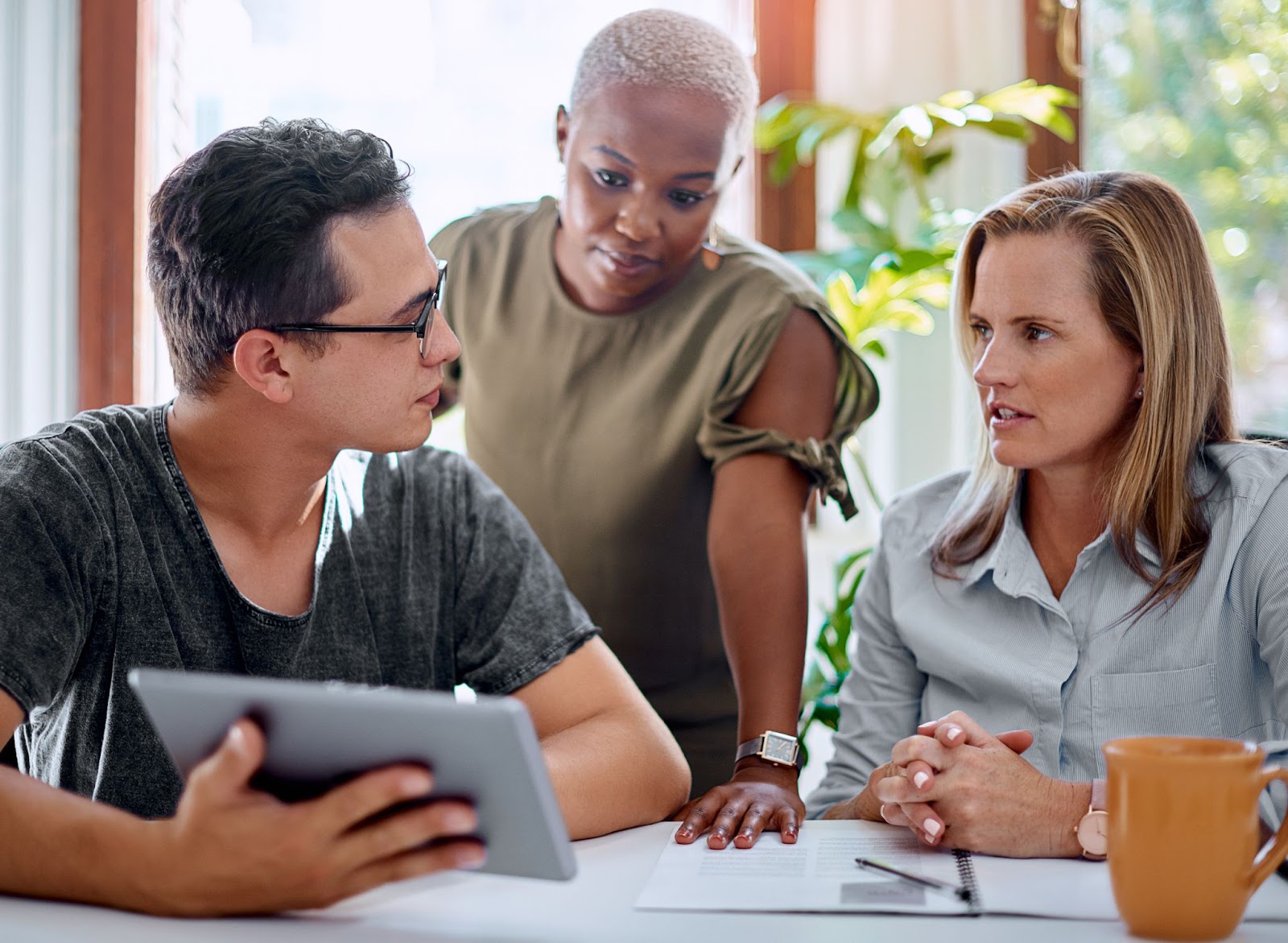 A male owner in a gray shirt presents data on a digital tablet, seemingly asking a question to his female consultants during a strategic meeting.